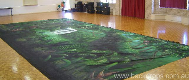 A large printed fabric backdrop with a jungle scene is laid out on a polished wooden floor in a spacious hall, creating the perfect setting for a fantastic time. In the background, there are stacked black cases and red curtains.