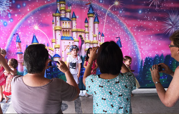 Adults take photos of children posing in front of a colorful fairy tale castle mural, enhanced by Party Drops, with fireworks lighting up the background.