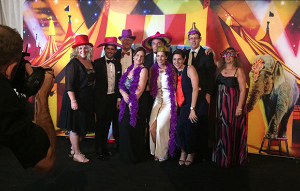 A lively group dressed in formal attire and colorful hats pose for a photo in front of a circus-themed backdrop, reminiscent of a grand party, while a skilled photographer captures the moment.
