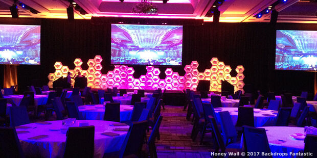 The stage, with its hexagonal backdrop and large screens, dominates the banquet hall filled with round tables and chairs. The modular design allows for versatility, while the lighting casts a purple and pink ambiance.