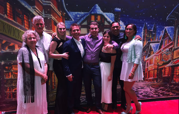 A group of people standing on a red carpet in front of a festive backdrop featuring a snowy village scene, celebrating at a holiday party. They are dressed in semi-formal attire and are smiling for the photo.