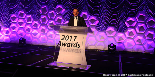 A man in a tuxedo stands at a clear podium labeled "2017 Awards" on a stage with modular purple geometric wall decor.