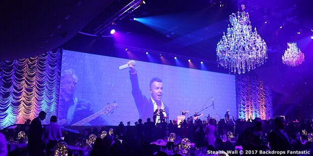 A singer on stage holds a microphone while performing at an event with modular purple lighting, chandeliers, and an audience seated at tables.