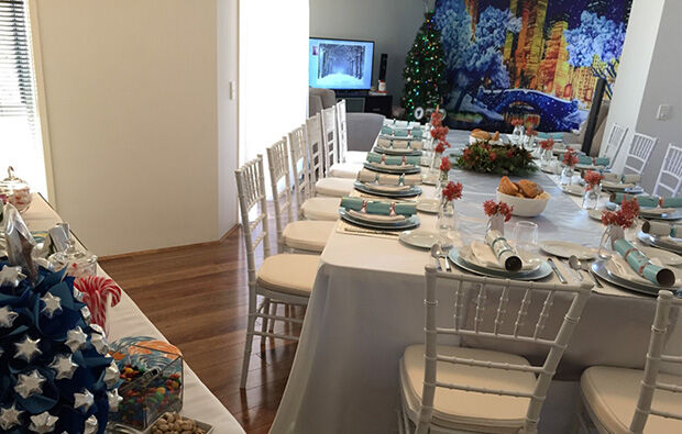 A long dining table with white chairs is set for a meal in a room with wooden floors. A decorated Christmas tree and a television displaying a festive scene add to the scenic backdrop.