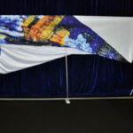 A person on a stepladder adjusts a large fabric banner with a cityscape and abstract design, draped over a metal frame, creating an impressive scenic backdrop.