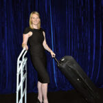 A person in a black dress stands beside a luggage cart while holding the handle of a suitcase, set against a scenic backdrop of deep blue curtains.