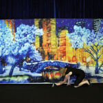 A person adjusts the bottom of a scenic backdrop displaying a winter cityscape with snow-covered trees and buildings against a night sky.