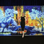 A person in a black dress poses in front of a scenic winter-themed backdrop, featuring a cityscape with snow-covered trees and buildings.
