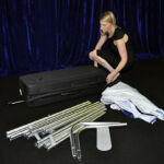 A woman kneels next to a suitcase, assembling metal poles and fabric pieces on a black floor, creating a scenic backdrop with dark blue curtains in the background.