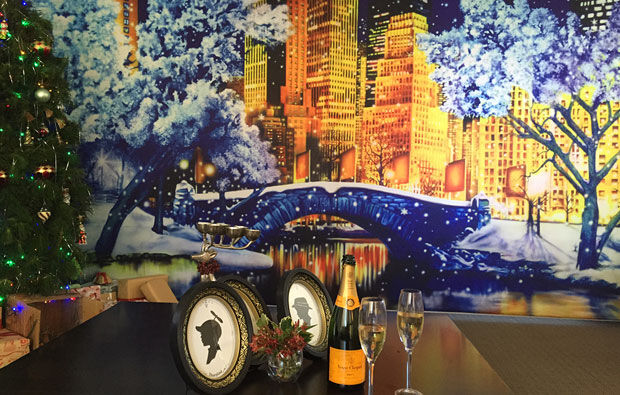 Festive table setup with champagne, two glasses, and framed silhouettes elegantly arranged in front of a scenic backdrop featuring a winter cityscape mural and a beautifully decorated Christmas tree.