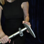 Person in a black dress holding a metal rod with a flat rectangular attachment against a scenic backdrop.