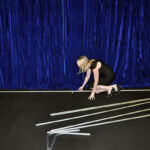 A woman in a black dress and heels kneels on the floor, assembling metal rods in front of a blue curtain, creating an elegant scenic backdrop next to a step ladder.
