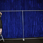 A person stands on a stepladder, adjusting a long metal rod with a base frame in front of a blue scenic backdrop.