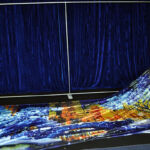 A person in black attire is laying out a large, colorful printed fabric on a floor, framed by a scenic blue curtain backdrop.