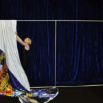 A person is setting up a display with a large printed fabric showing a cityscape, hanging it over a simple metal frame against a dark blue curtain background, creating the perfect scenic backdrop.