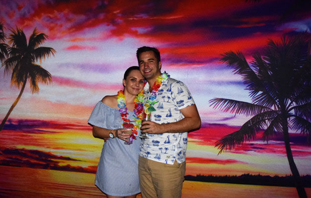 A couple stands together in front of a vibrant tropical sunset backdrop, wearing leis and holding drinks from Party Drops. Palm trees are silhouetted against the colorful sky.