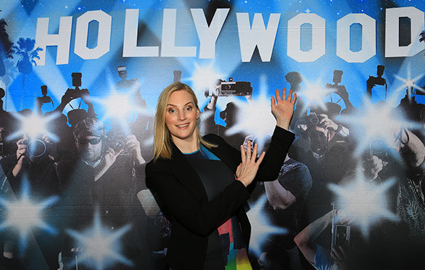 A woman posing happily in front of a background featuring the word "Hollywood" and an image of paparazzi taking pictures, giving off major party drops vibes.