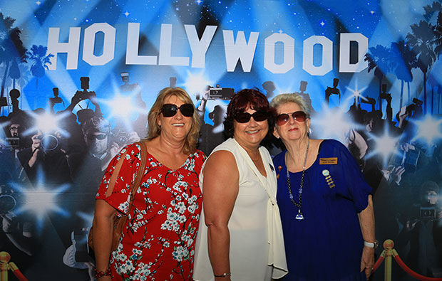 Three women wearing sunglasses pose in front of a Hollywood-themed backdrop featuring photographers and spotlights, enjoying the lively energy of the party.