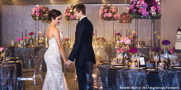 A couple in formal attire holds hands in an elegantly decorated room with modular tables set for an event, adorned with flowers and candles.