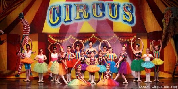 Performers in colorful costumes dance energetically on stage in front of a large "CIRCUS" sign with vibrant decorations, creating a dynamic and mesmerizing slider view.