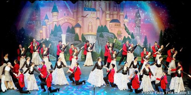 A group of performers in red jackets and elaborate dresses are on stage in front of a castle backdrop, engaged in a synchronized slider dance routine.