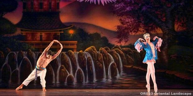 Two ballet dancers gracefully perform on stage against a backdrop depicting an oriental landscape with a waterfall and traditional architecture. The dancer in blue glides effortlessly, while the shirtless performer executes a flawless slider, enhancing their mesmerizing dance.