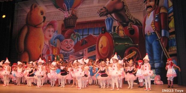 Children in various costumes dance on stage in front of a backdrop featuring toy characters, including a bear, clown, and toy soldier.