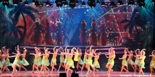 A group of dancers in colorful costumes slide gracefully across the stage, surrounded by a tropical-themed background and vibrant lighting.