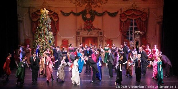 A large group of people in Victorian-era attire dance in a lavishly decorated parlor, with a large Christmas tree, an ornate fireplace, and a festive centerpiece featuring a holiday slider in the background.