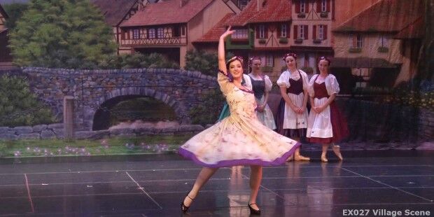 A dancer performs a ballet move on stage, gracefully sliding across the floor in front of a backdrop depicting a quaint village with a stone bridge. Three other dancers in traditional costumes observe from the background.